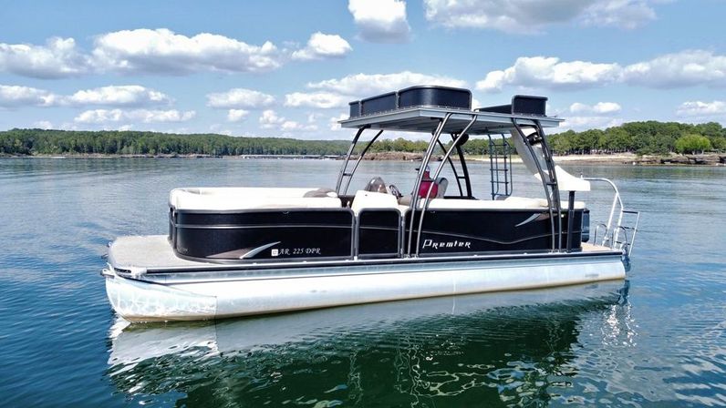Sleek black-and-white pontoon boat floating on a glassy lake under a sunny blue sky with puffy clouds and a tree-lined shoreline