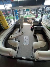Pontoon boat interior with curved beige-and-gray vinyl seating, carpeted deck, removable cup-holder table and forward helm, docked in a covered marina slip with green lake water and neighboring boats visible.
