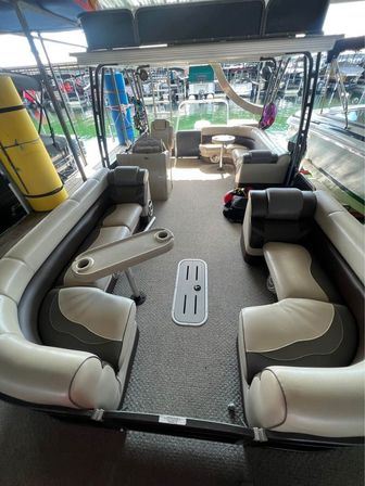 Pontoon boat interior with curved beige-and-gray vinyl seating, carpeted deck, removable cup-holder table and forward helm, docked in a covered marina slip with green lake water and neighboring boats visible.