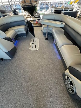 Cozy pontoon boat interior at a marina with gray carpeted deck, curved light- and dark-gray vinyl bench seating, a small removable table with cup holders, built-in marine speakers and blue LED accent lights, view toward the docked bow.