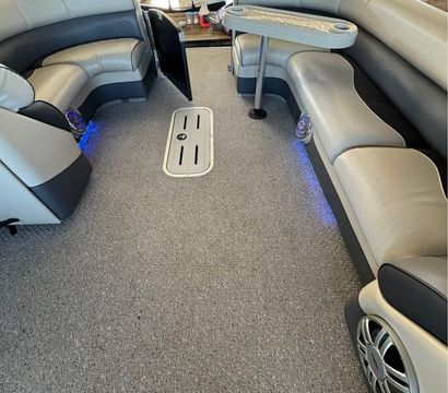 Cozy pontoon boat interior at a marina with gray carpeted deck, curved light- and dark-gray vinyl bench seating, a small removable table with cup holders, built-in marine speakers and blue LED accent lights, view toward the docked bow.
