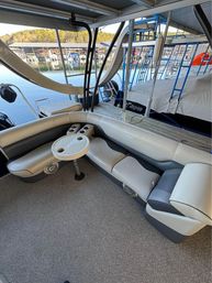 Pontoon boat interior with curved beige and gray cushioned seating, round table with cupholders, docked at a covered marina on a calm lake with tree-lined shore.