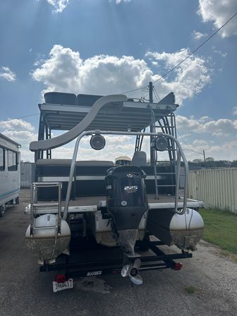 Rear view of a pontoon boat on a trailer with a Yamaha 225 outboard, twin aluminum pontoons, curved waterslide and wake tower with mounted speakers, parked on a gravel lot under a bright blue sky with fluffy clouds.