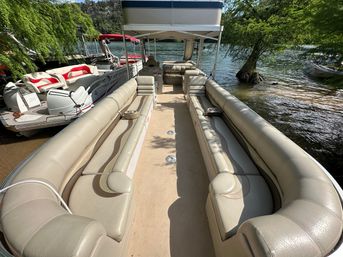 Sunlit beige pontoon boat interior with long cushioned bench seats, cup holders and canopy, docked on a leafy lake shoreline with calm water.