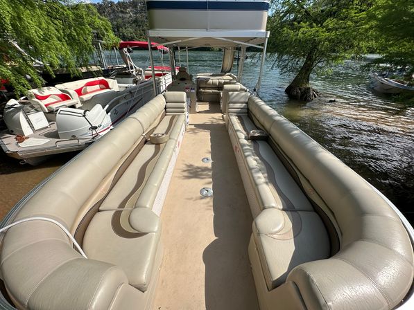 Sunlit beige pontoon boat interior with long cushioned bench seats, cup holders and canopy, docked on a leafy lake shoreline with calm water.