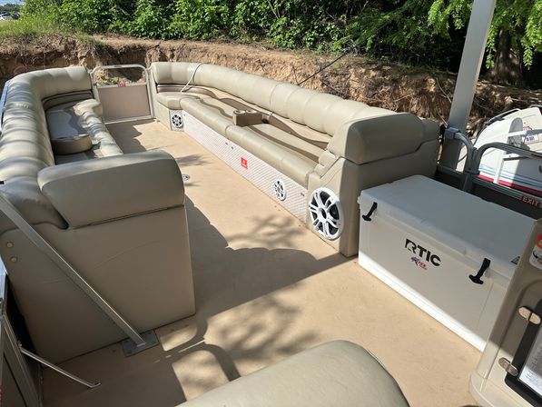 Beige pontoon boat deck with wraparound cushioned seating, white marine speakers, a large white cooler, and cupholders, moored near a leafy lakeshore.