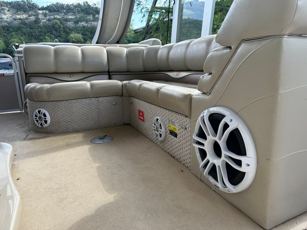 Beige L-shaped vinyl seating on a pontoon boat with white marine speakers and textured panels, lake shoreline visible in the background