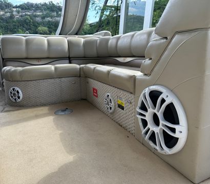 Beige L-shaped vinyl seating on a pontoon boat with white marine speakers and textured panels, lake shoreline visible in the background