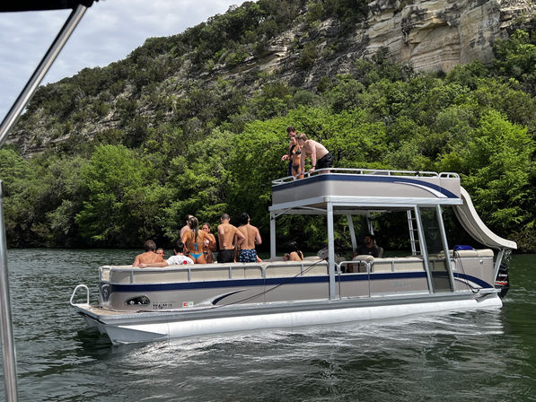 Pontoon boat with upper deck and water slide carrying a group of people enjoying a summer boat party on a green, tree-lined lake beside rocky cliffs
