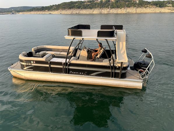 Pontoon boat with cushioned seating, upper sun deck and aft slide, Yamaha outboard and a person at the helm cruising on a calm lake near a rocky shoreline at golden hour
