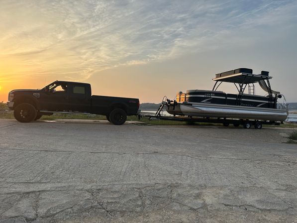 Black pickup truck towing a pontoon boat on a trailer at a lakeside boat ramp during sunset with orange sky and calm water