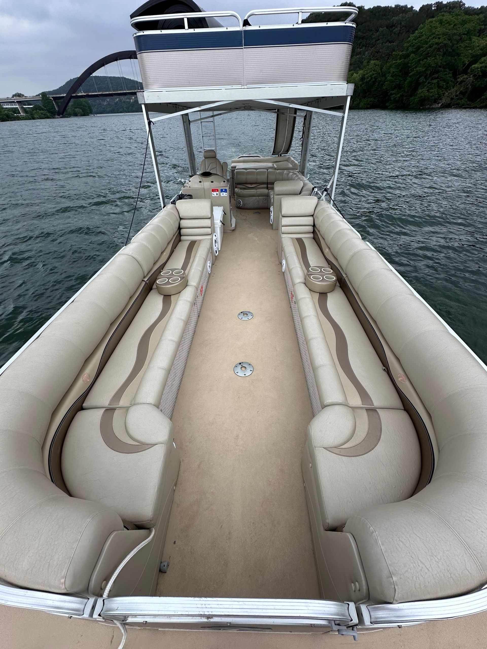 Spacious beige pontoon boat interior with long center aisle, cushioned bench seats and cup holders, anchored on a lake with an arched bridge and tree-lined shore under a cloudy sky