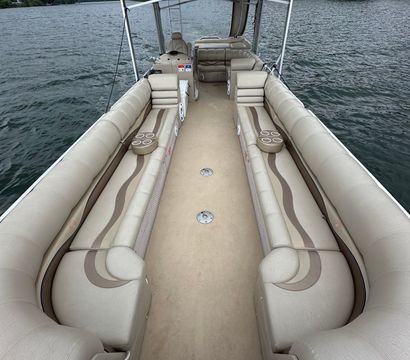 Spacious beige pontoon boat interior with long center aisle, cushioned bench seats and cup holders, anchored on a lake with an arched bridge and tree-lined shore under a cloudy sky