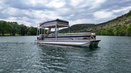 Pontoon boat floating on a calm lake with tree-lined shoreline and rocky hills under a dramatic cloudy sky