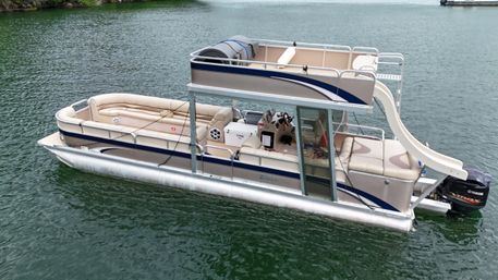 Two-level beige pontoon party boat with upper deck and water slide, Yamaha outboard, floating on a green lake