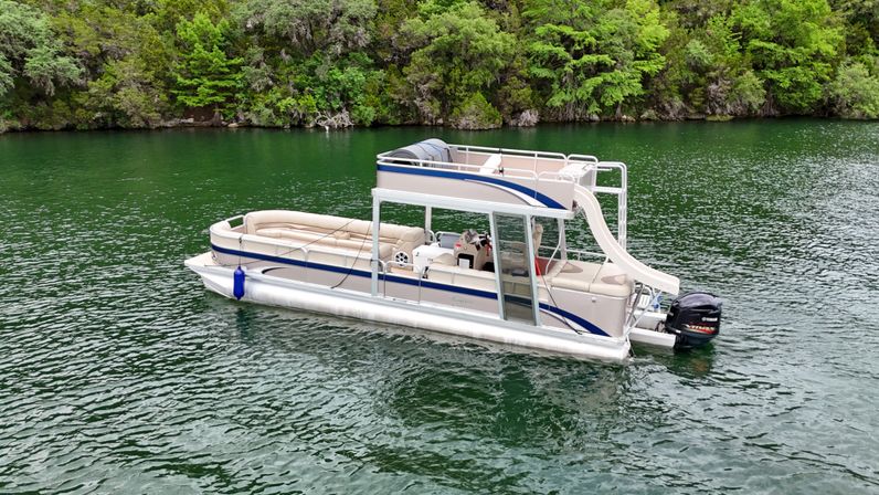 Pontoon boat with upper deck and slide floating on emerald-green lake beside a dense, forested shoreline
