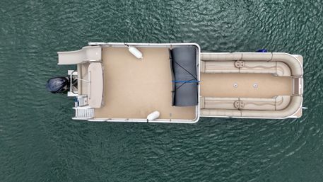 Aerial top-down view of a beige pontoon boat floating on green lake water, featuring wraparound cushioned seating, swim platform with outboard motor, a deck slide and a folded black canopy.
