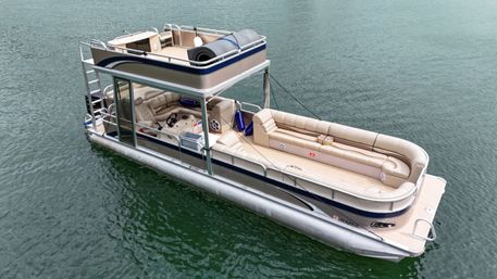 Aerial view of a two-level beige and navy pontoon boat with wraparound tan lounge seating and upper deck, floating on green lake water