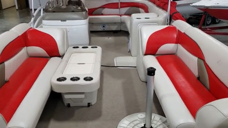 Pontoon boat interior with red-and-white vinyl bench seating, center table/cooler with multiple cup holders, carpeted open deck and helm in the background.