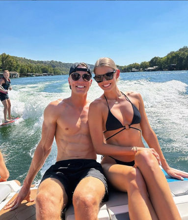 Sun-soaked young couple in sunglasses and swimwear sitting on a speedboat swim platform on a blue lake, with a wakesurfer riding the boat’s wake and tree-lined shoreline behind them.