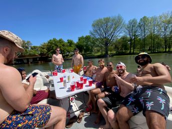 Group of friends on a sunny pontoon boat party at a calm lake—shirtless men gathered around a table with red solo cups and drinks, trees lining the shoreline under a clear blue sky.