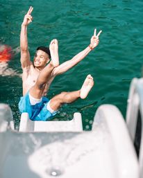Shirtless man in blue swim trunks mid-air off a boat slide into turquoise water, smiling and flashing peace signs — fun summer jump