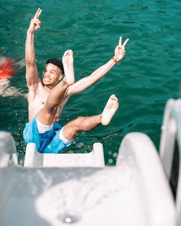 Shirtless man in blue swim trunks mid-air off a boat slide into turquoise water, smiling and flashing peace signs — fun summer jump