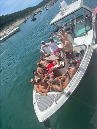 Group of men relaxing on a white motorboat anchored in clear blue lake water near a tree-lined shore, enjoying a sunny summer boat party.