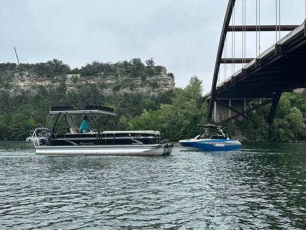 Two recreational boats — a black pontoon and a blue motorboat — float on a calm lake beneath a steel arch bridge, with tree-covered limestone cliffs and an overcast sky.