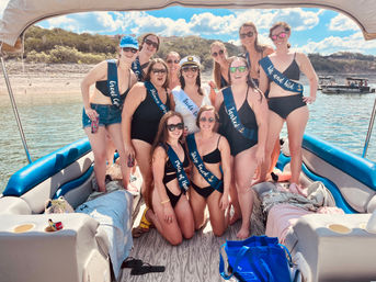 Smiling bachelorette group in black swimsuits and sashes posing on a pontoon boat at a sunny lake shoreline