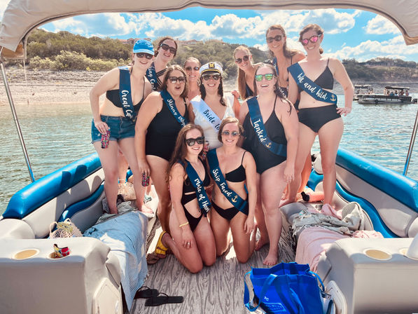 Smiling bachelorette group in black swimsuits and sashes posing on a pontoon boat at a sunny lake shoreline