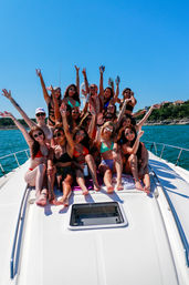 Group of young women in colorful bikinis cheering and raising drinks on the bow of a white yacht, sunny summer day on clear turquoise water with a tree-lined shoreline in the background.