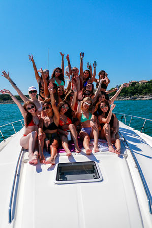 Group of young women in colorful bikinis cheering and raising drinks on the bow of a white yacht, sunny summer day on clear turquoise water with a tree-lined shoreline in the background.