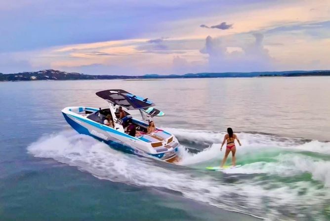 Wake surfer riding a green-tinted wake behind a blue-and-white motorboat on a calm lake at sunset, passengers watching from the boat.