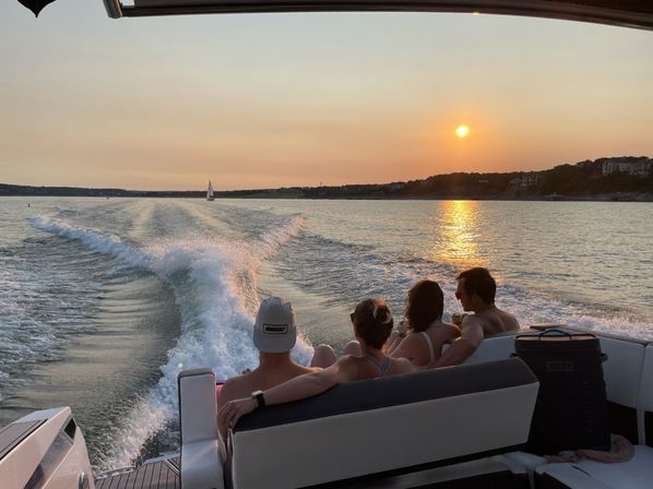 Four people relaxing on the stern of a motorboat at sunset, watching a golden lake with the boat's wake stretching back and a small sailboat on the horizon.