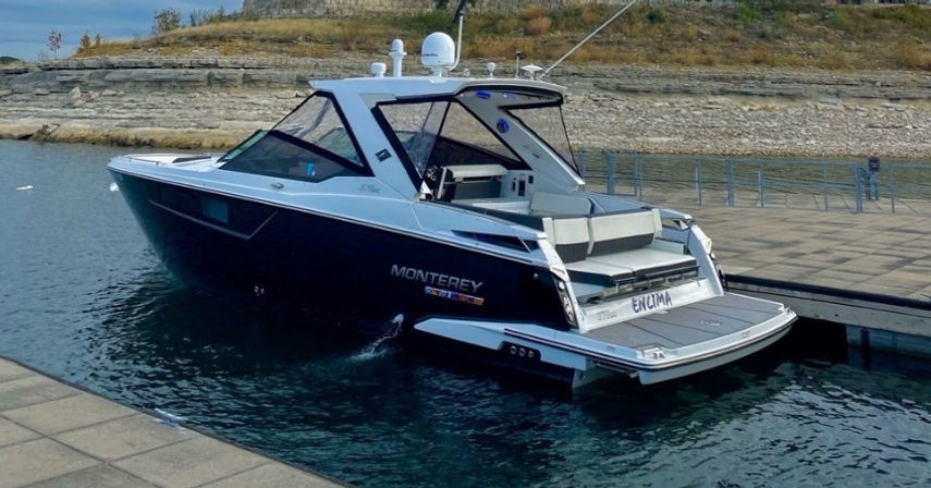Sleek black-and-white luxury motorboat docked at a concrete marina, showing open swim platform, cushioned aft seating and calm water with a rocky shoreline in the background.