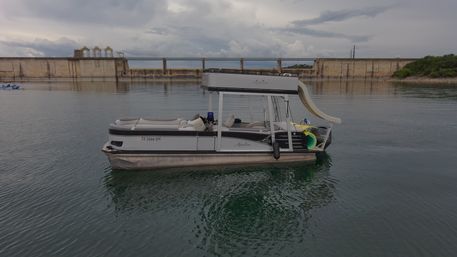 Pontoon party boat with rooftop waterslide floating on a calm reservoir near a concrete dam under a moody cloudy sky