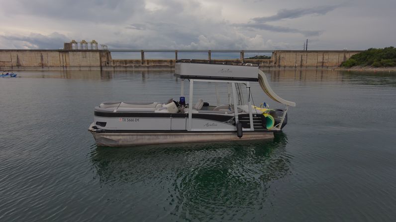 Pontoon party boat with rooftop waterslide floating on a calm reservoir near a concrete dam under a moody cloudy sky