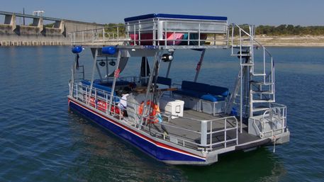 Two-level aluminum pontoon boat with blue seating, a spiral rear ladder and deck cooler floating on a calm lake near a concrete dam and rocky shoreline under a clear sky
