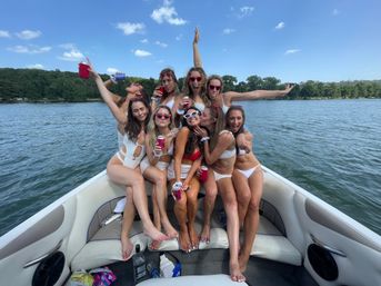 Group of friends in swimsuits on the bow of a pontoon boat at a sunlit lake, laughing and posing with sunglasses and canned drinks against a backdrop of trees and blue sky