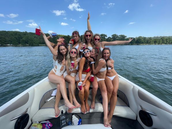 Group of friends in swimsuits on the bow of a pontoon boat at a sunlit lake, laughing and posing with sunglasses and canned drinks against a backdrop of trees and blue sky