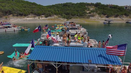 Summer lake party on a Texas lake with a crowded flotilla of pontoon boats and colorful float mats, people in swimsuits, American and Texas flags, and a green tree-lined shoreline.