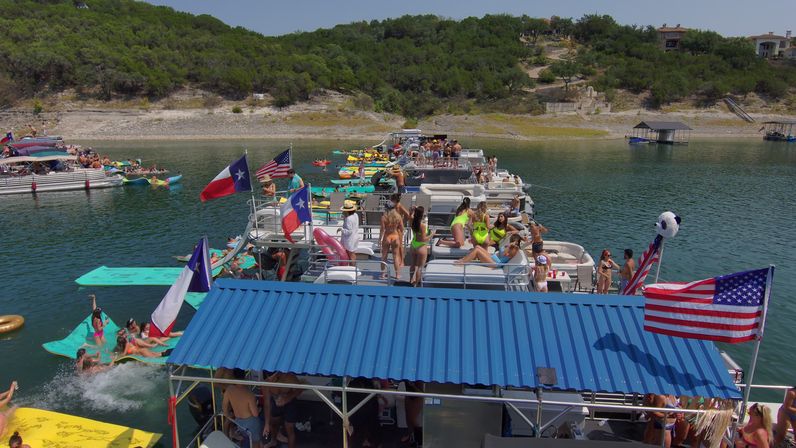 Summer lake party on a Texas lake with a crowded flotilla of pontoon boats and colorful float mats, people in swimsuits, American and Texas flags, and a green tree-lined shoreline.
