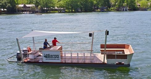 Wooden-deck pontoon party boat with shaded canopy and two people at the helm, drifting on a sunny lake with tree-lined residential shoreline in the background.