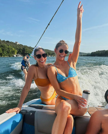 Two women in bikinis and sunglasses smiling on the back of a speedboat during a sunny lake day — one in a blue patterned bikini raises an arm and holds a travel tumbler while the other in a mustard bikini laughs, a wakeboarder rides the boat’s wake and tree-lined shoreline stretches in the background.