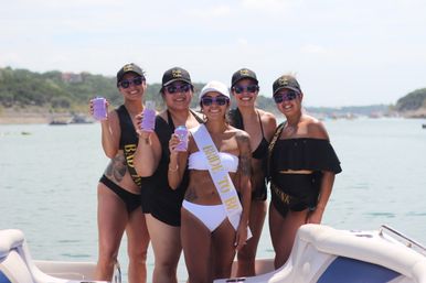 Five women on a boat at a lake celebrating a bachelorette party, bride-to-be in a white sash and bikini with friends in black swimwear and matching caps holding drinks, sunny shoreline in the background.