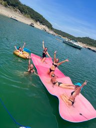 Sunny lake day: five women in bikinis cheering and waving on a giant pink floating mat and a gold inflatable tube, with boats and a tree-lined shore in the background.