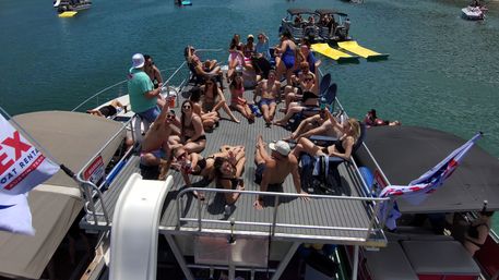 Sun-soaked pontoon boat party on a clear lake, dozens of young adults in swimsuits lounging, sunbathing and waving on the upper deck with a small slide, nearby boats and floating docks visible