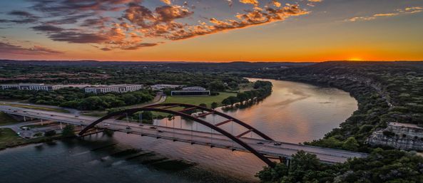 Aerial sunset view of Pennybacker Bridge (360 Bridge) spanning the Colorado River in Austin, Texas, with vibrant orange sky and water reflections.