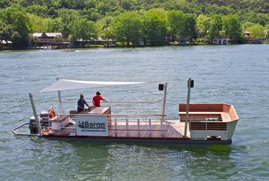 Sunlit wooden party pontoon boat with white canopy and two people aboard cruising on a tree-lined lake in front of waterfront homes.